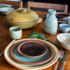 A styled tabletop arrangement of handmade ceramic tableware on a wooden dining table, including stacked plates, bowls, cups, a vase, and a lidded casserole dish, with a sprig of rosemary in the foreground.
