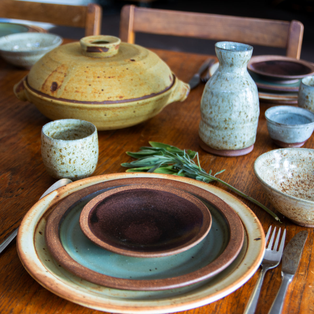 A styled tabletop arrangement of handmade ceramic tableware on a wooden dining table, including stacked plates, bowls, cups, a vase, and a lidded casserole dish, with a sprig of rosemary in the foreground.